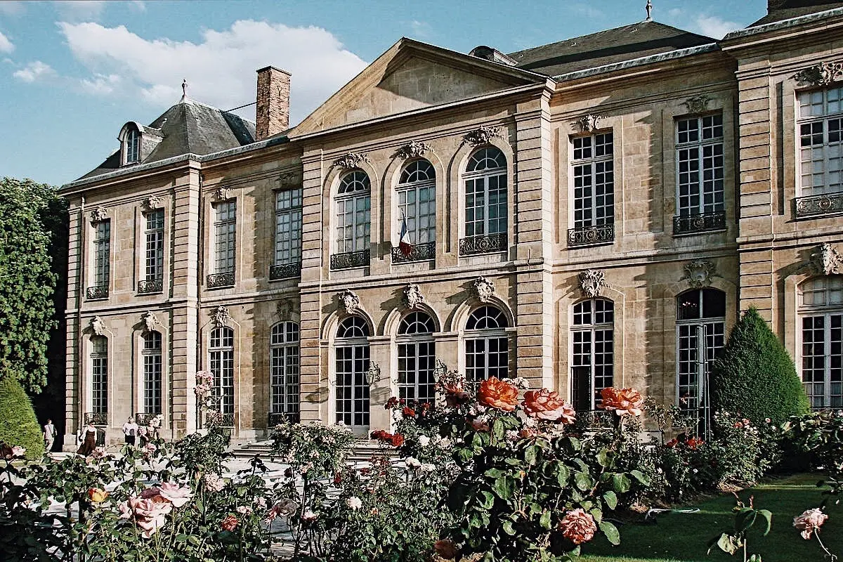 Un bâtiment historique en pierre avec des fenêtres cintrées, entouré de rosiers en fleurs sous un ciel bleu.