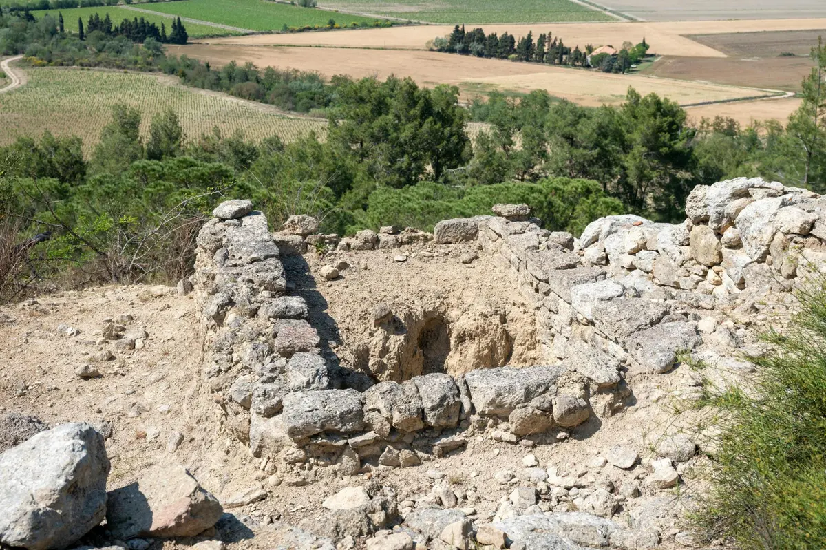 Ruines d'une petite structure en pierre avec un trou au centre, entourée d'un paysage rural avec des champs et des arbres.