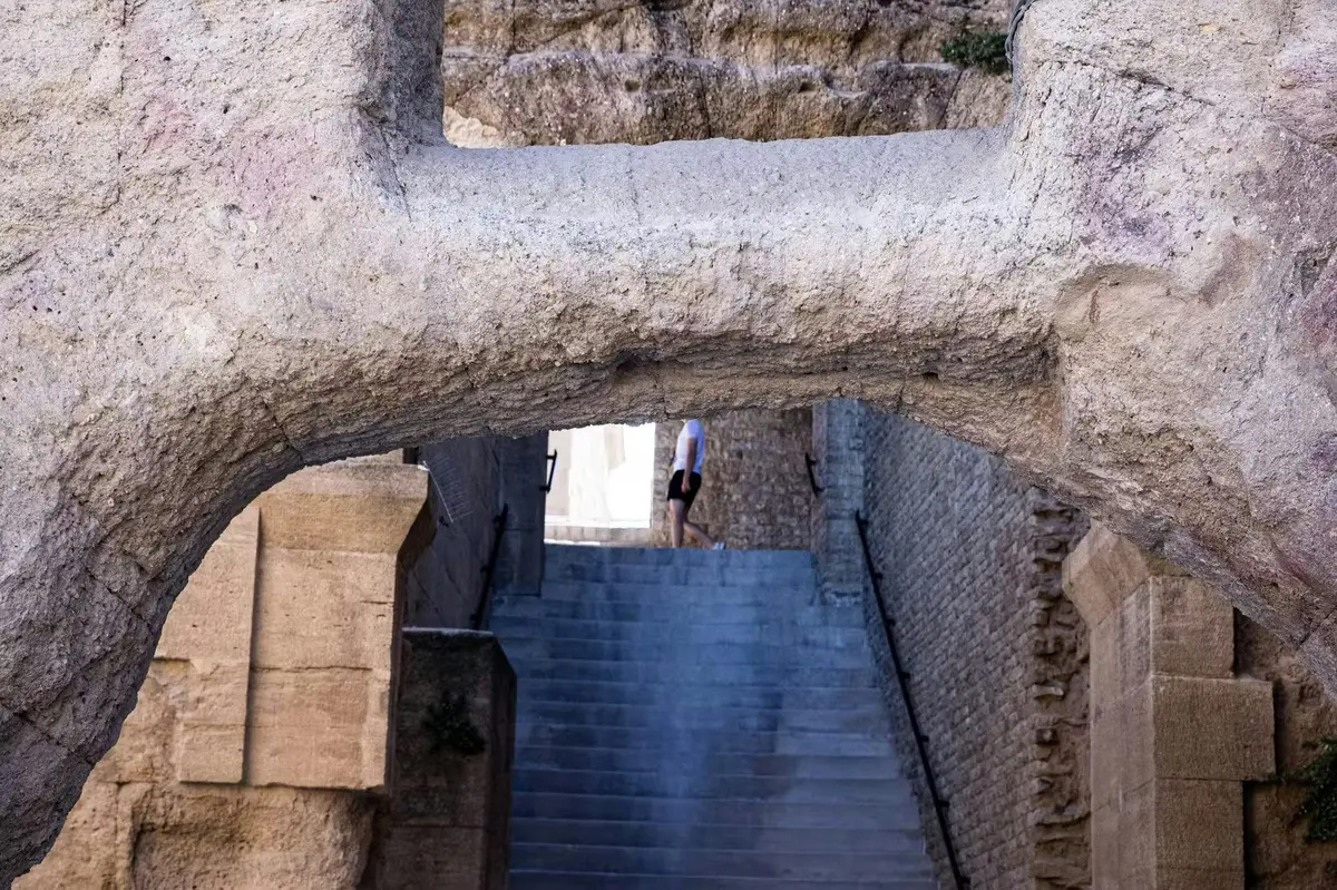 Une personne en short monte un long escalier de pierre sous une grande arche de pierre, avec des murs anciens en briques des deux côtés.