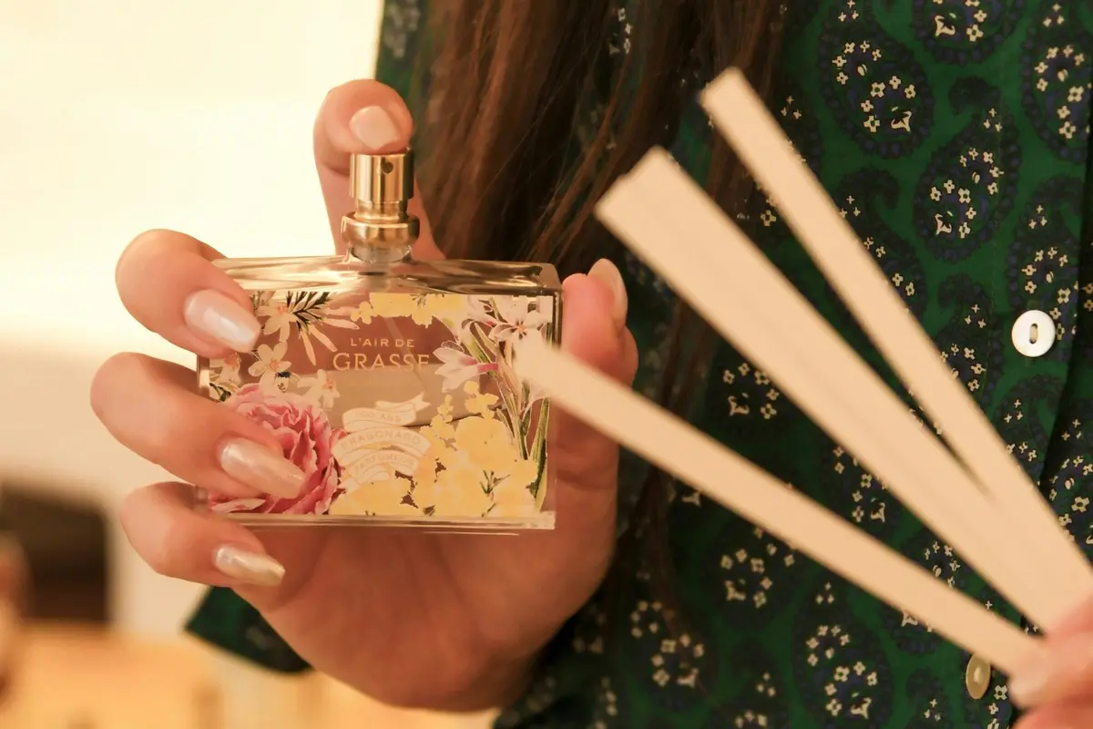 Une personne tenant un flacon de parfum orné d'un motif floral et portant l'étiquette « L'air de Grasse », avec des bâtonnets parfumés en bois au premier plan.