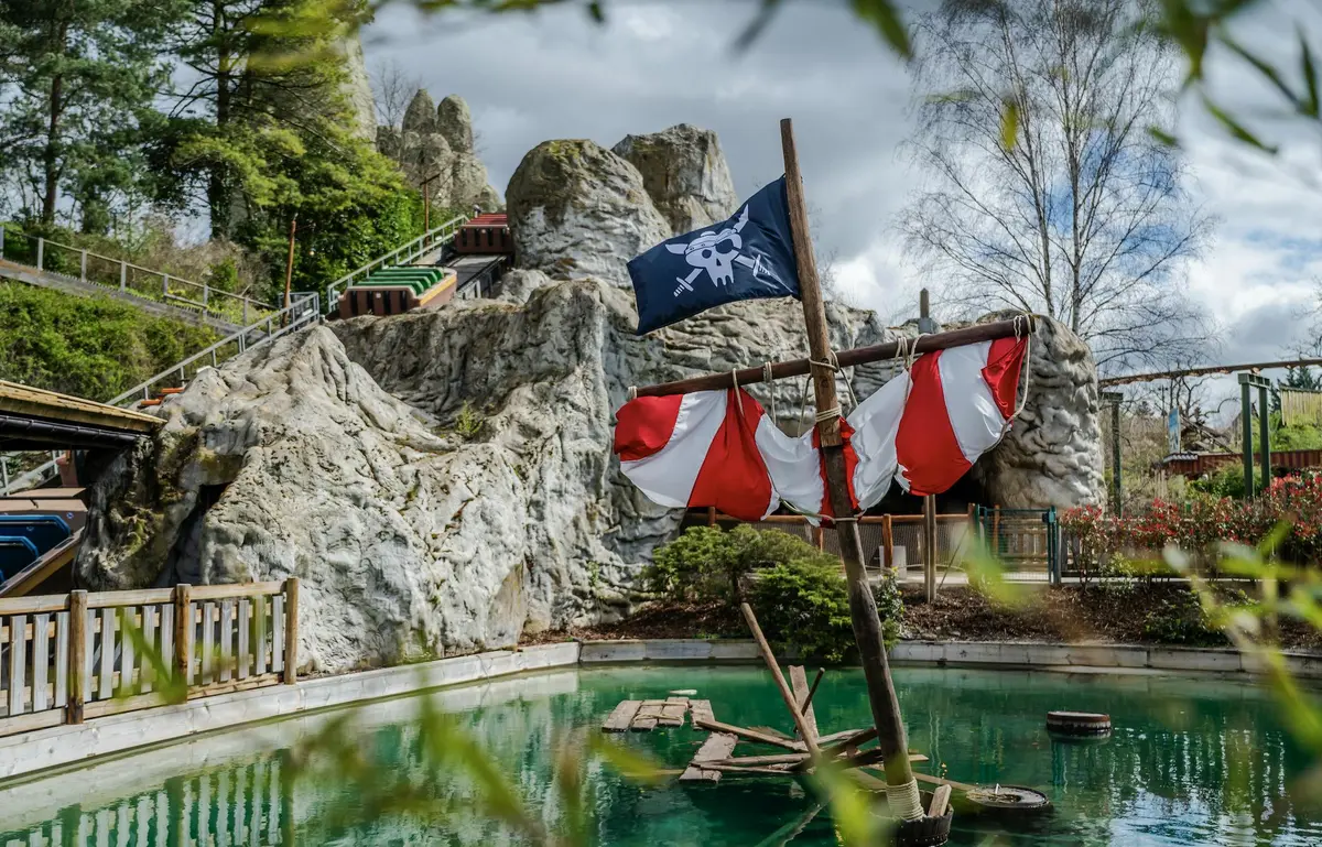 A pirate ship mast with red and white sails and a black skull flag in a pond, surrounded by rocks and greenery.