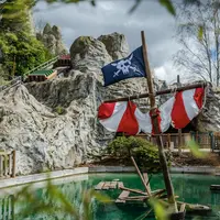 A pirate ship mast with red and white sails and a black skull flag in a pond, surrounded by rocks and greenery. &copy; @sylvaincambon