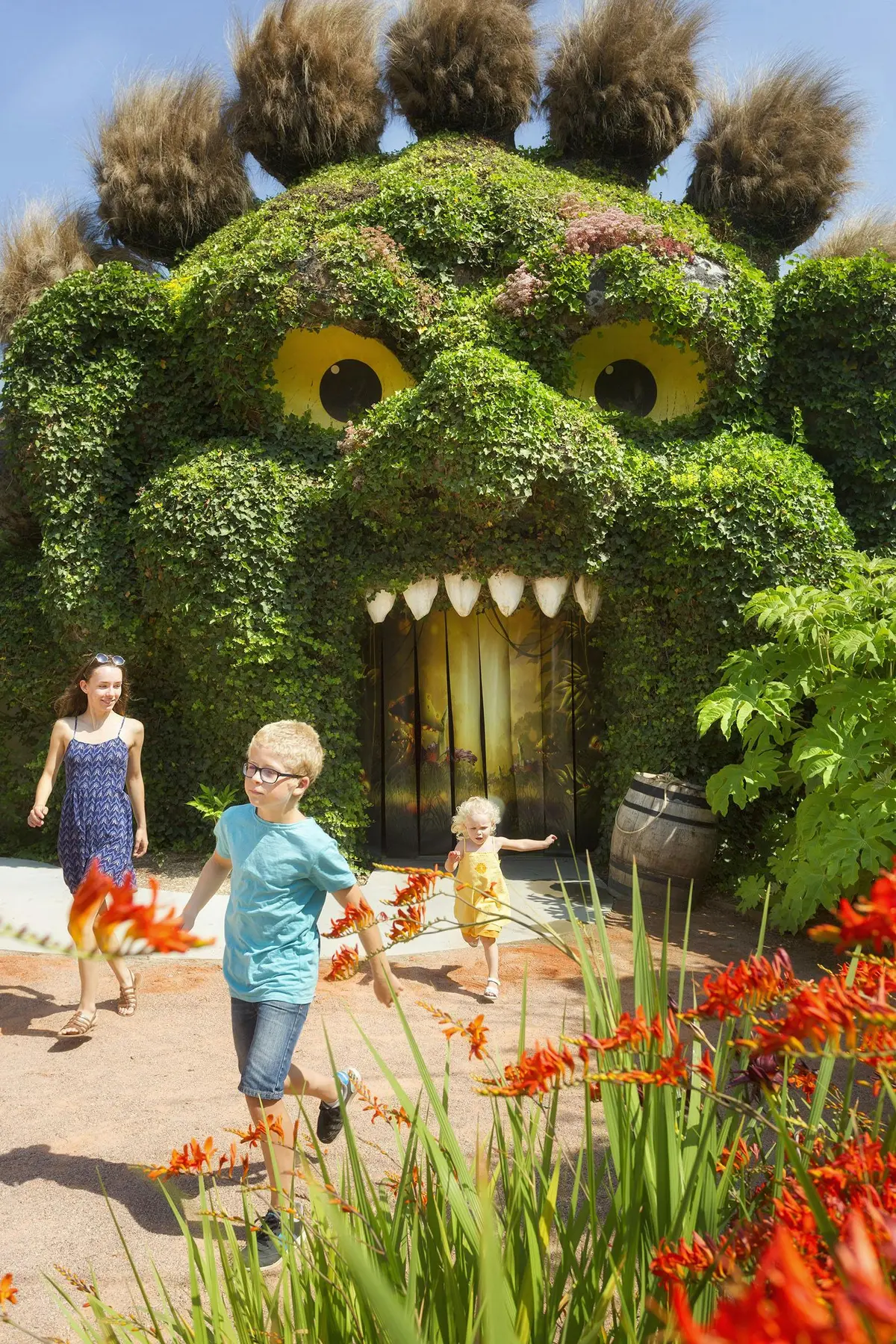 Deux enfants et un adulte marchant devant une grande structure végétale avec des yeux et une bouche. Des fleurs rouges au premier plan.