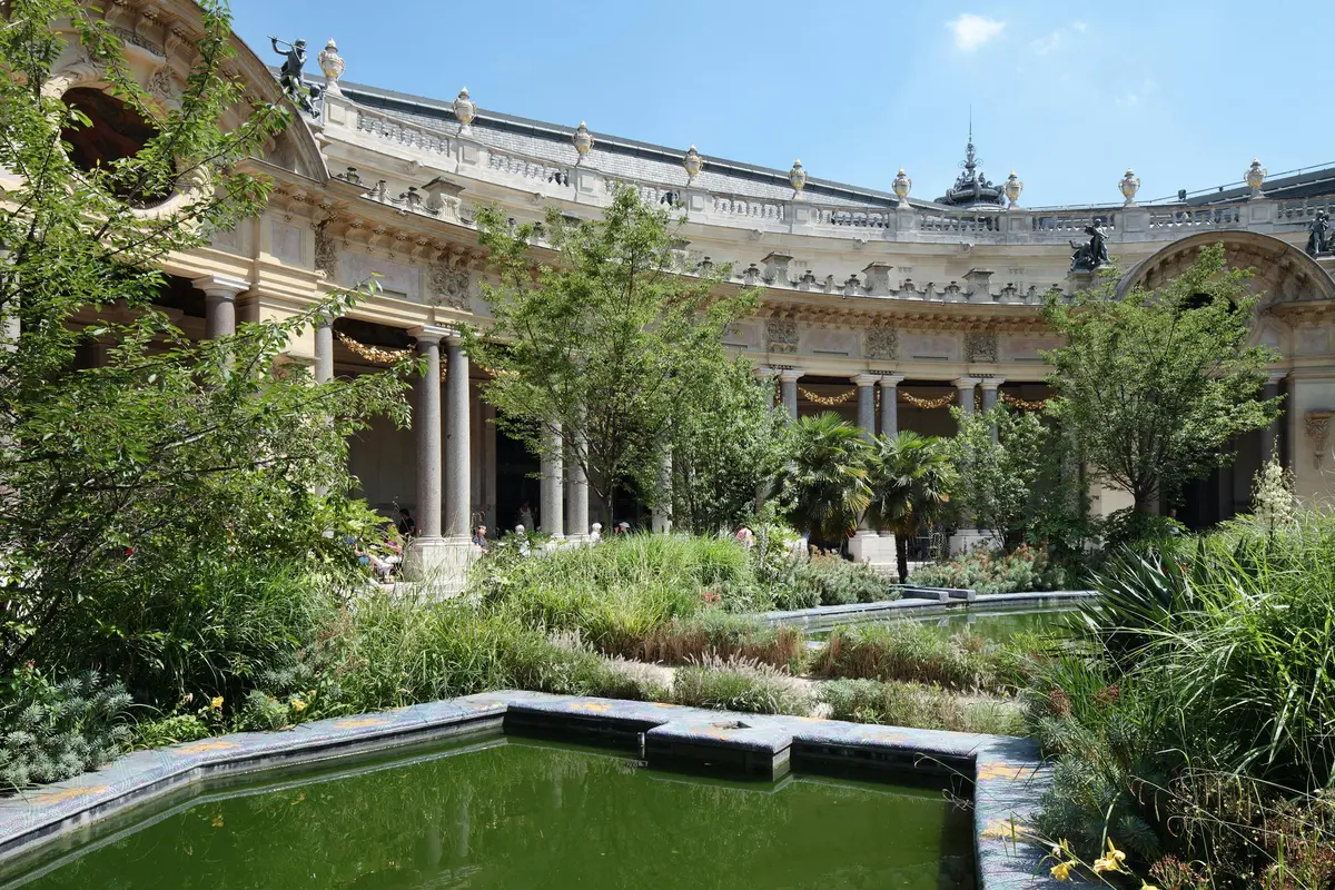 A historic building with colonnades and statues, surrounded by lush greenery and a rectangular pond in the foreground.