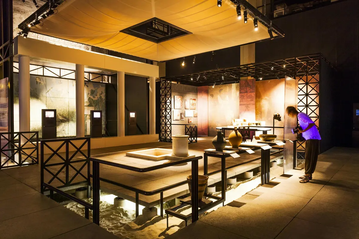 Exposition de poteries et d'objets anciens dans une salle bien éclairée avec des grilles en bois et un sol illuminé.