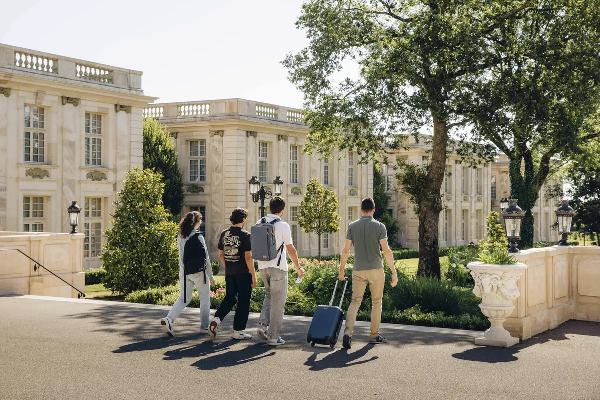 Quatre personnes marchant vers un bâtiment de style classique avec des bagages, entouré d'arbres et de verdure par une journée ensoleillée.