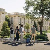 Quatre personnes marchant vers un bâtiment de style classique avec des bagages, entouré d'arbres et de verdure par une journée ensoleillée. DR