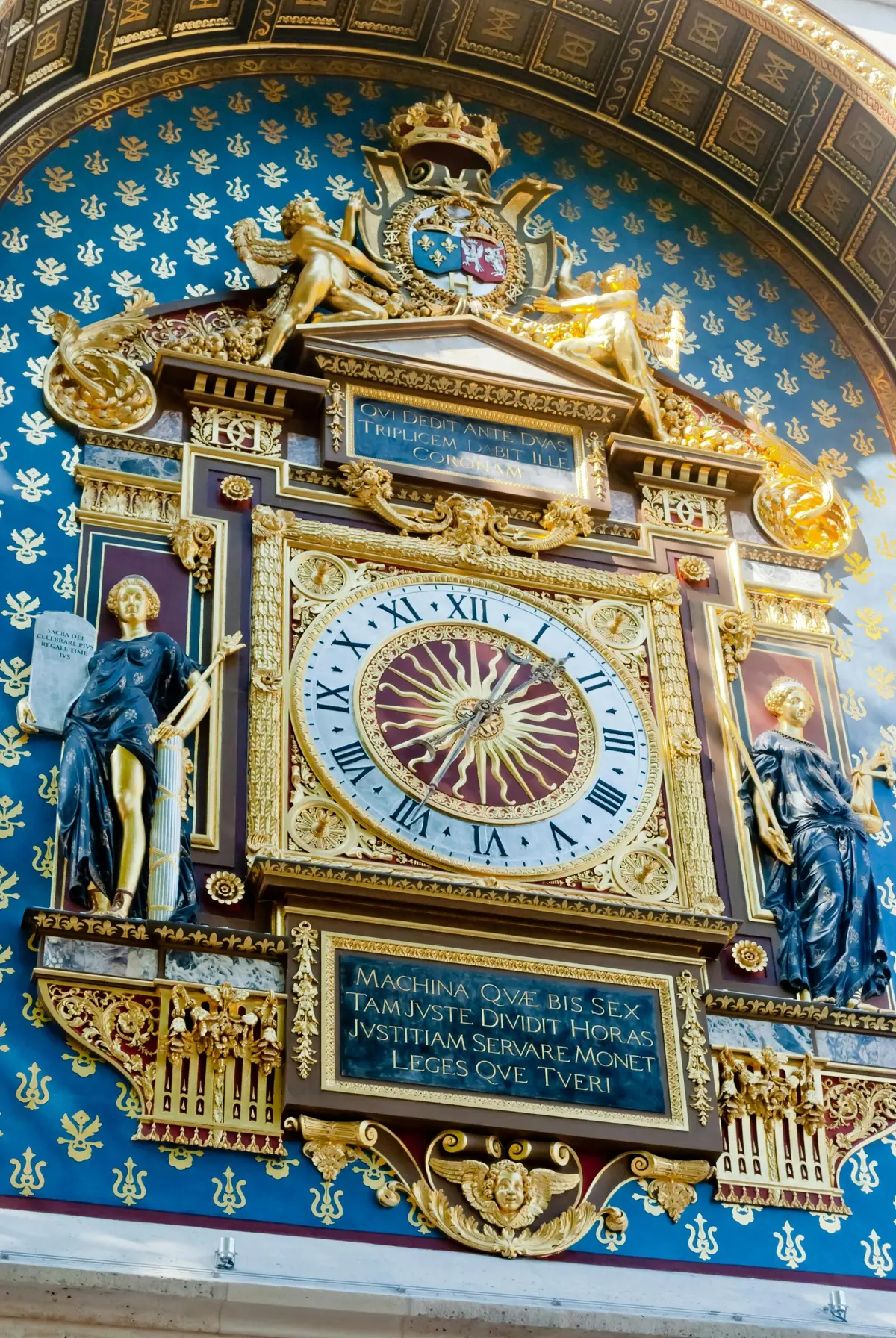 Une horloge richement décorée avec des chiffres romains, flanquée de deux statues, d'éléments dorés et d'inscriptions en latin.