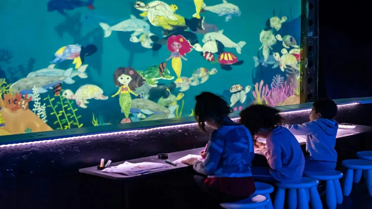 Deux enfants colorient à une table devant un mur sur le thème de l'océan, couvert de dessins de sirènes, de poissons et de créatures marines.