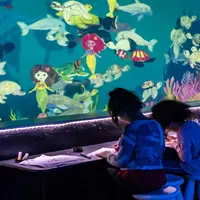 Deux enfants colorient à une table devant un mur sur le thème de l'océan, couvert de dessins de sirènes, de poissons et de créatures marines. DR