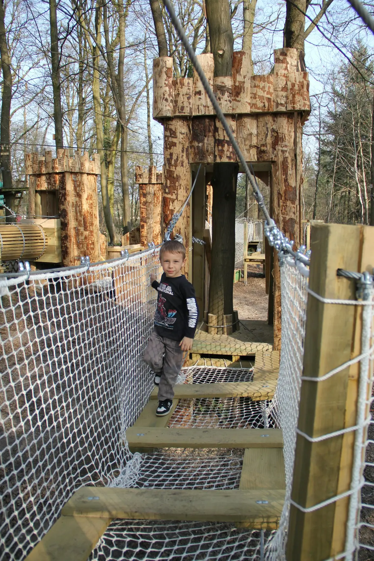 Un enfant marche sur un pont de corde dans une aire de jeux en bois entourée d'arbres.