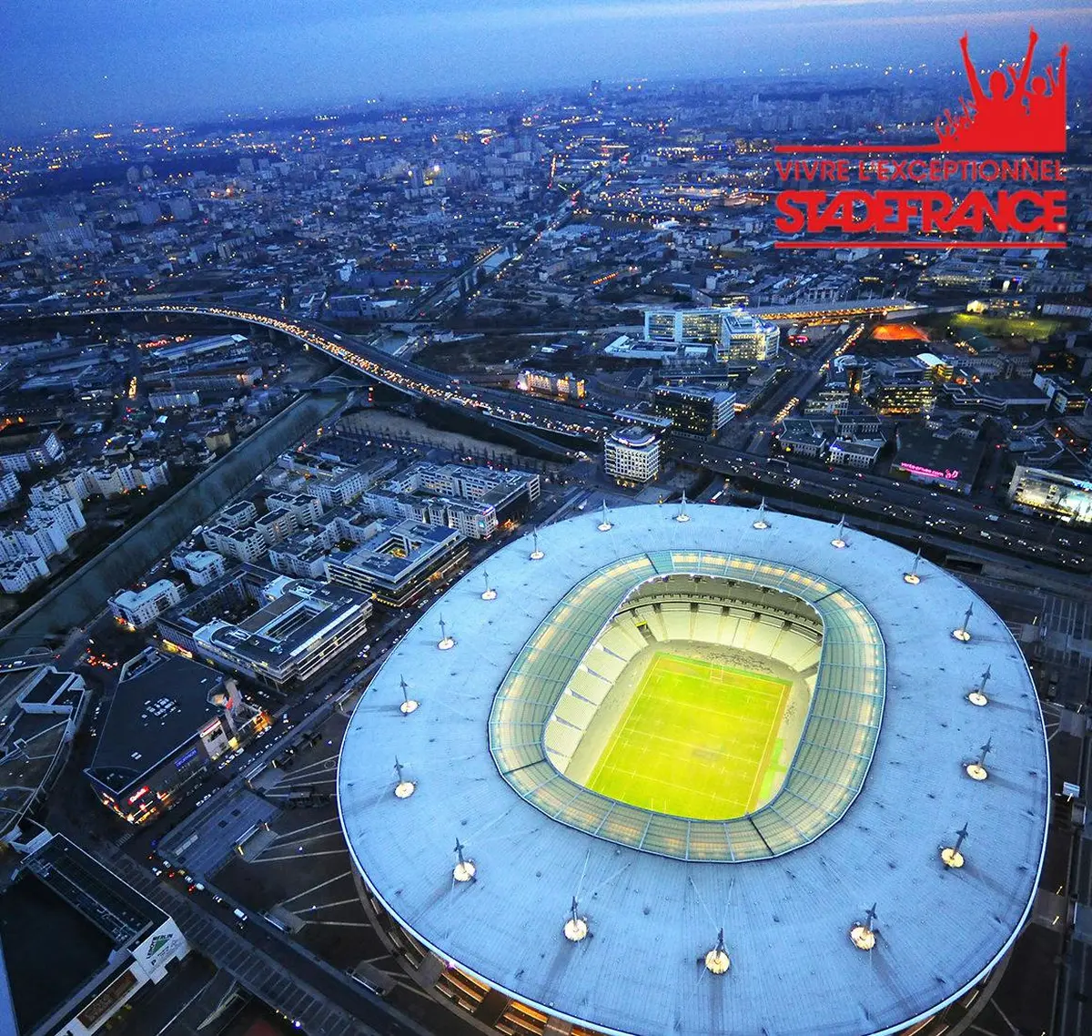 Vue aérienne d'un stade bien éclairé dans une ville animée au crépuscule, avec des routes, des bâtiments et une rivière visibles.