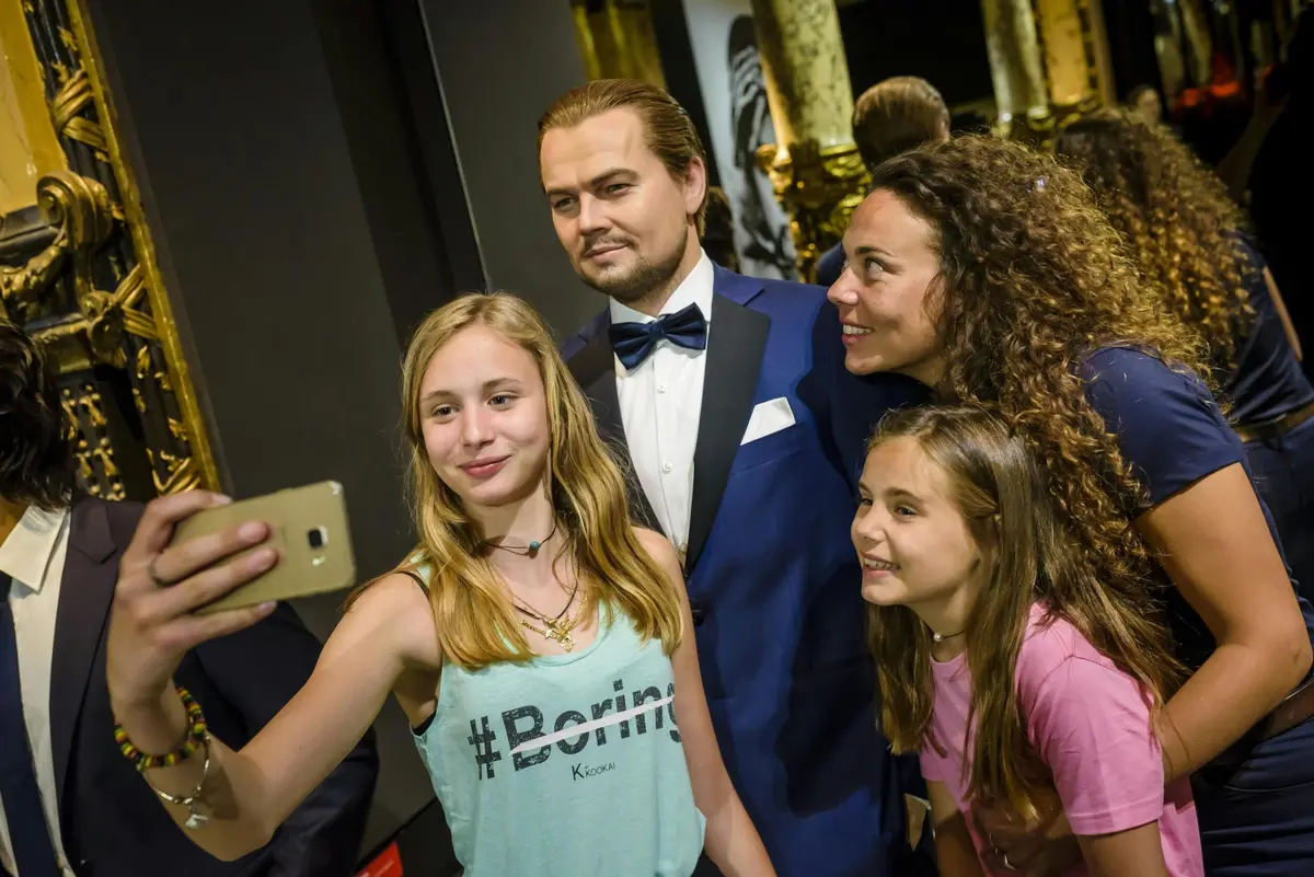 Trois filles prennent un selfie avec un personnage de cire en costume bleu dans un musée ; l'une d'entre elles porte un t-shirt Boring.
