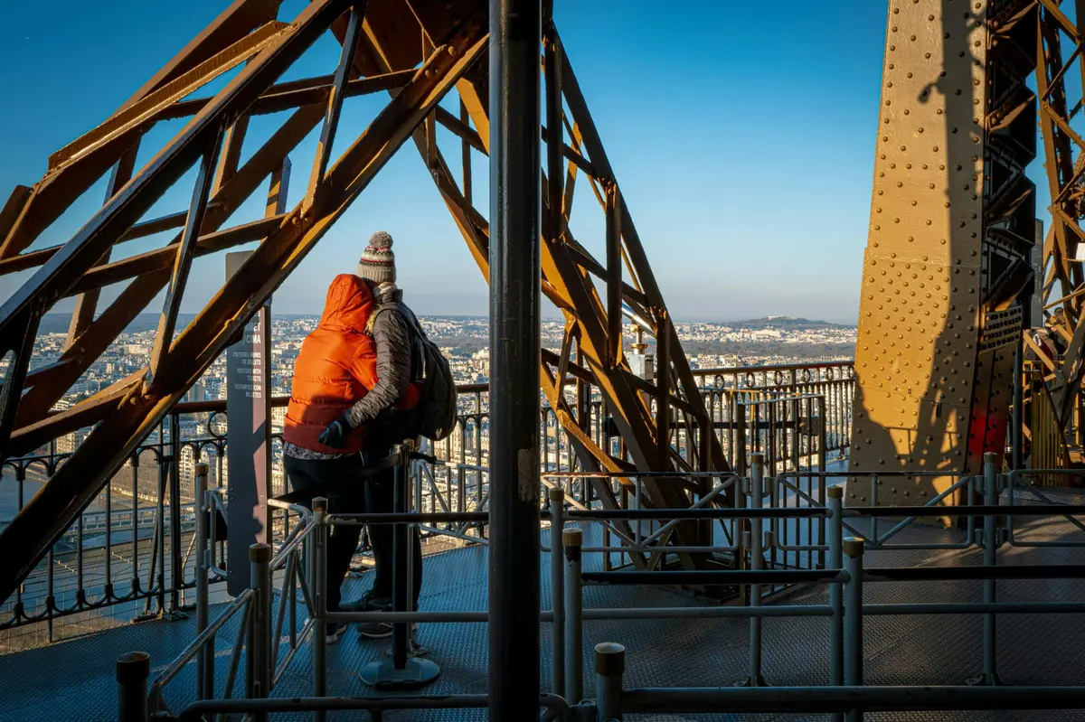 Deux personnes se tiennent sur une plate-forme de la Tour Eiffel, entourée de poutres métalliques et de garde-corps, surplombant un paysage urbain.
