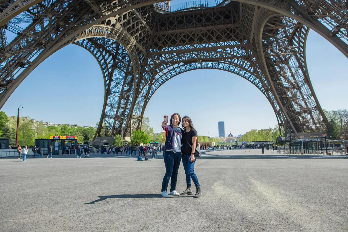 Deux personnes se tiennent au pied de la Tour Eiffel et prennent un selfie par une journée ensoleillée, sous un ciel dégagé.