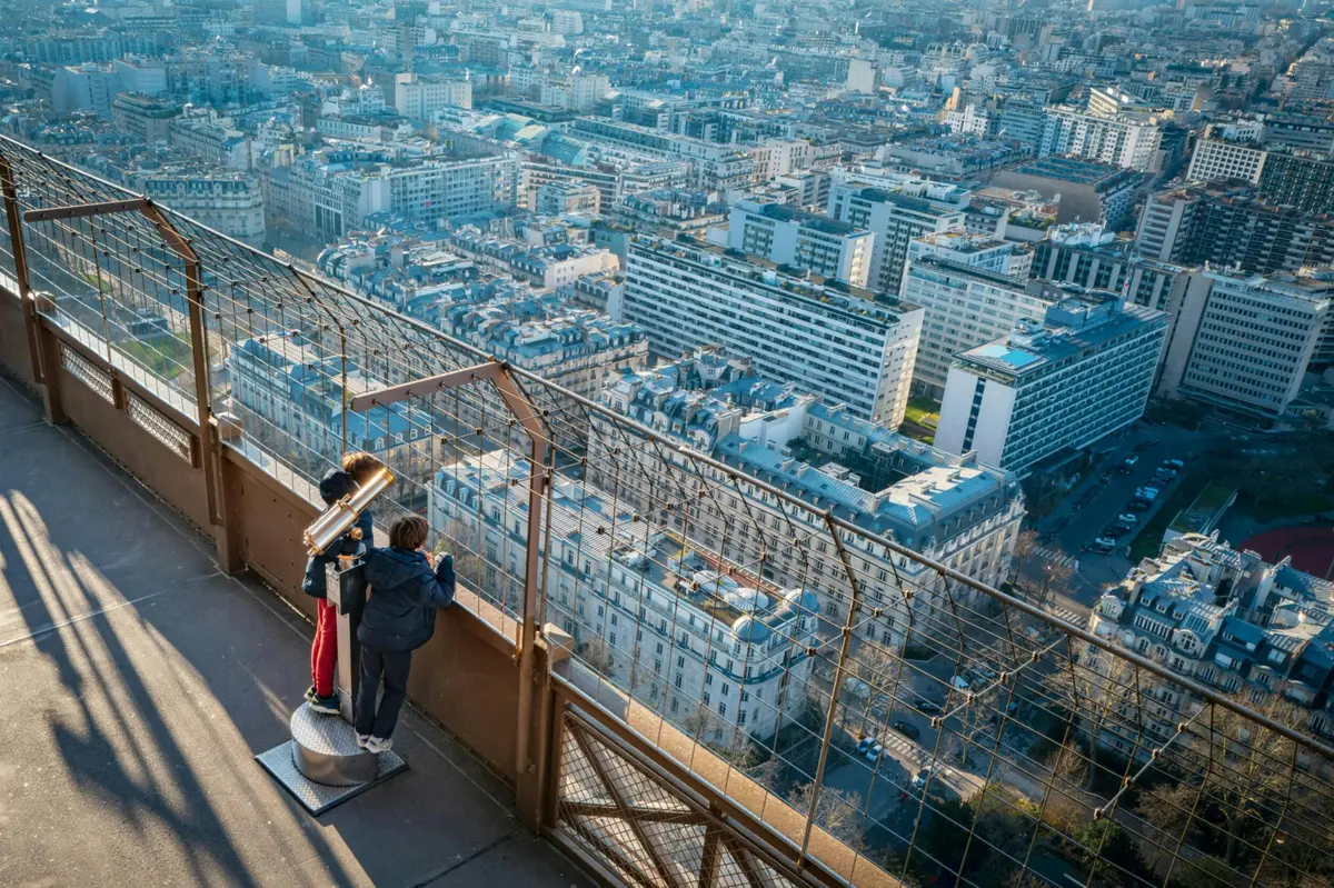 Deux personnes sur un pont d'observation regardant un paysage urbain à travers un télescope, avec une clôture métallique au premier plan.