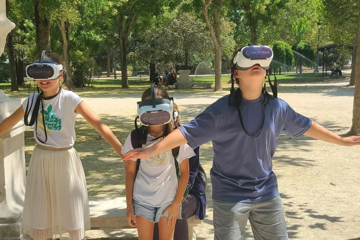 Trois personnes portant des casques de réalité virtuelle à l'extérieur, dans un parc ensoleillé, avec des arbres et des bancs en pierre en arrière-plan.