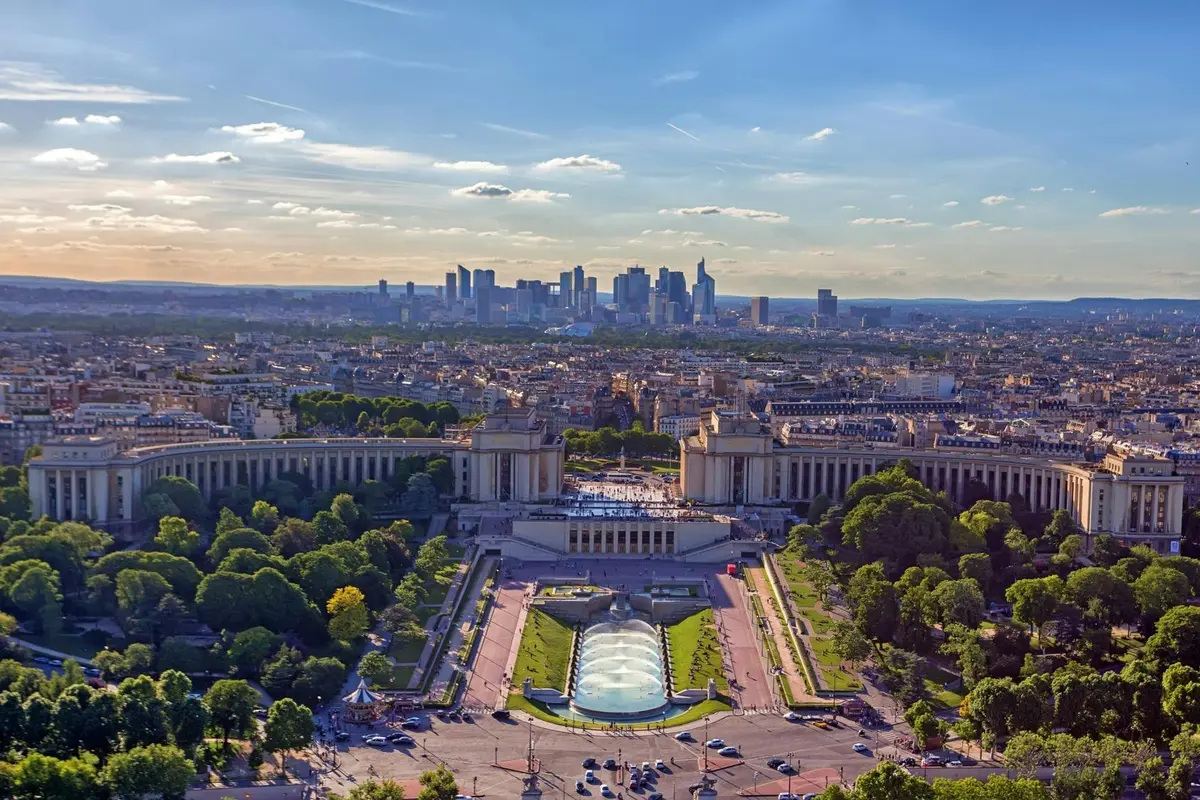 Vue plongeante sur les jardins du Trocadéro à Paris, avec leurs fontaines, leurs bâtiments symétriques et la silhouette de la ville au loin.