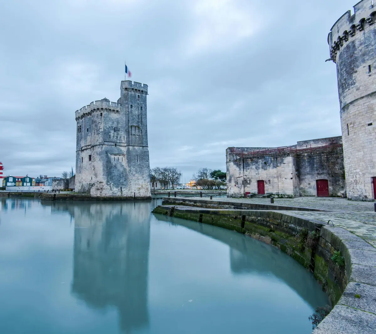 Une haute tour historique en pierre avec un drapeau se dresse au bord d'une eau calme, avec d'autres structures en pierre et un ciel nuageux à l'arrière-plan.