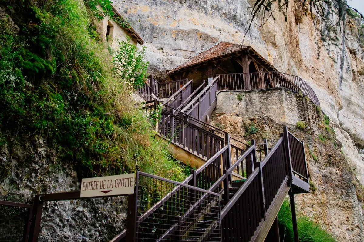 Des escaliers en bois mènent à un bâtiment rustique construit à flanc de falaise. Un panneau indique ENTREE DE LA GROTTE.