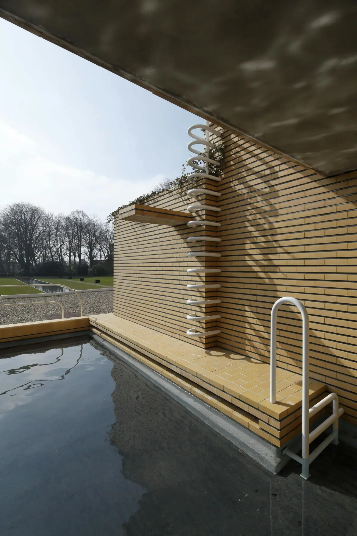 Piscine extérieure moderne avec des murs en briques, des mains courantes blanches et un escalier en colimaçon. Arbres et pelouse visibles en arrière-plan.