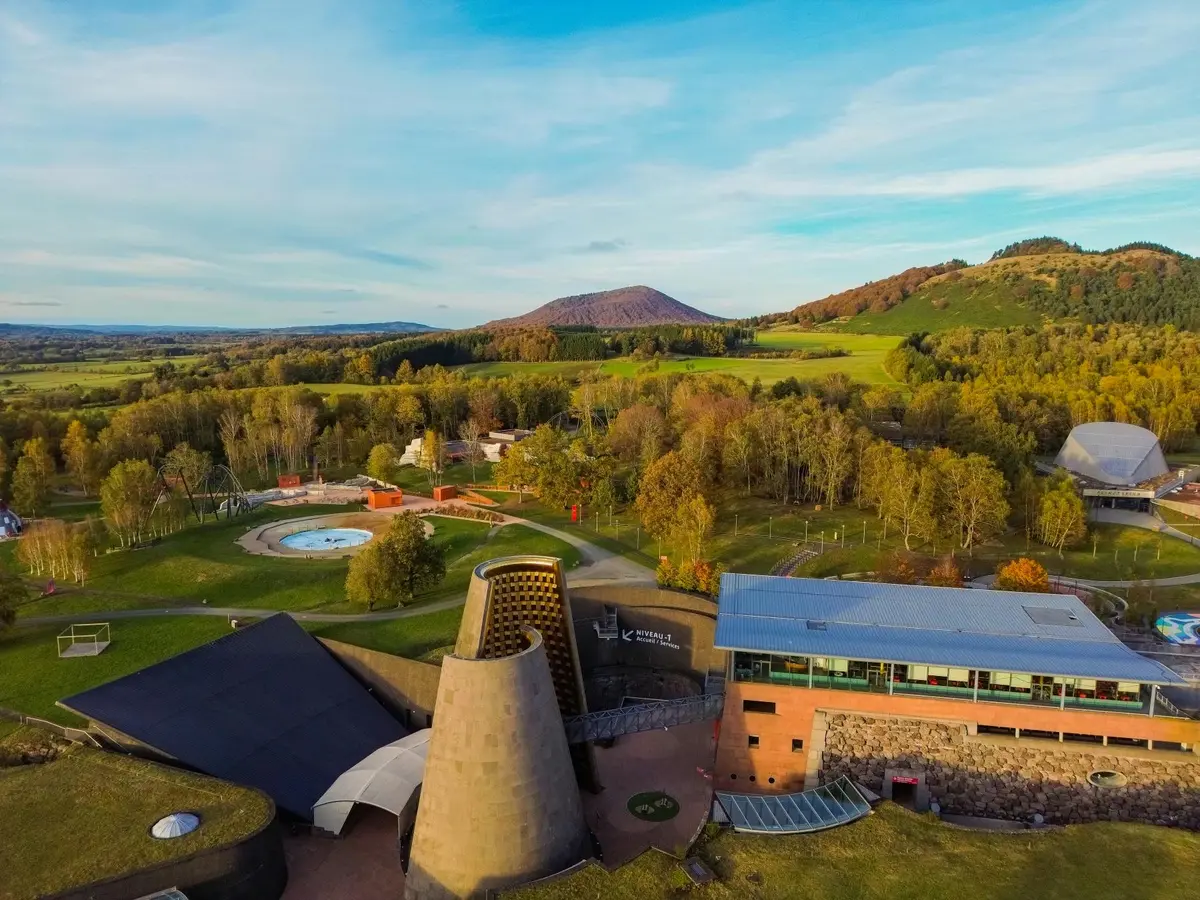 Vue aérienne d'un parc avec des bâtiments, une piscine, des arbres et une montagne en arrière-plan sous un ciel dégagé.