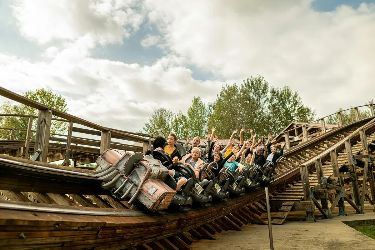 Des personnes montent sur des montagnes russes en bois, les bras levés, entourées d'arbres et d'un ciel nuageux.