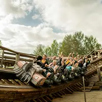 Des personnes montent sur des montagnes russes en bois, les bras levés, entourées d'arbres et d'un ciel nuageux. &copy; Walibi Rhone Alpes