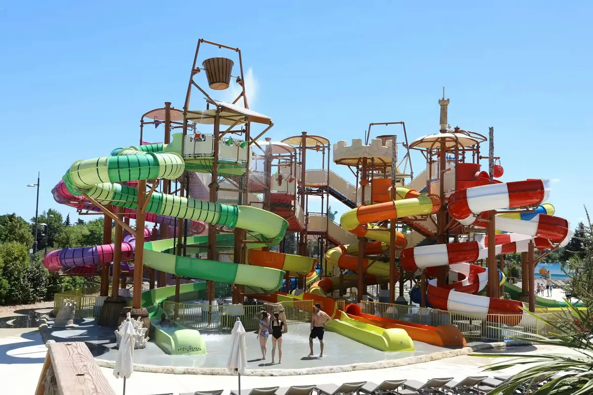 Un parc aquatique haut en couleur avec des toboggans, un seau basculant et des gens qui se promènent dans une pataugeoire sous un ciel bleu limpide.