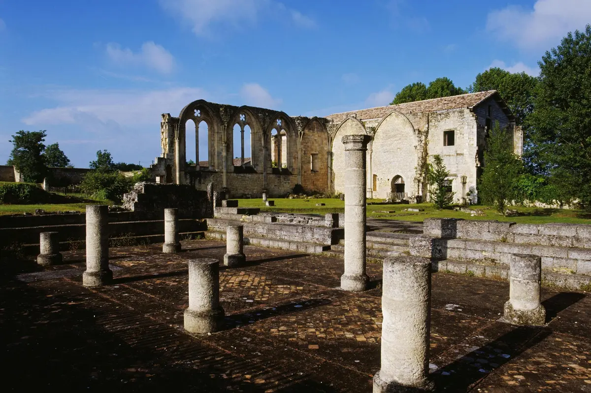 Ruines d'un vieux bâtiment en pierre avec de hautes colonnes et des fenêtres cintrées sous un ciel bleu clair. Arbres en arrière-plan.