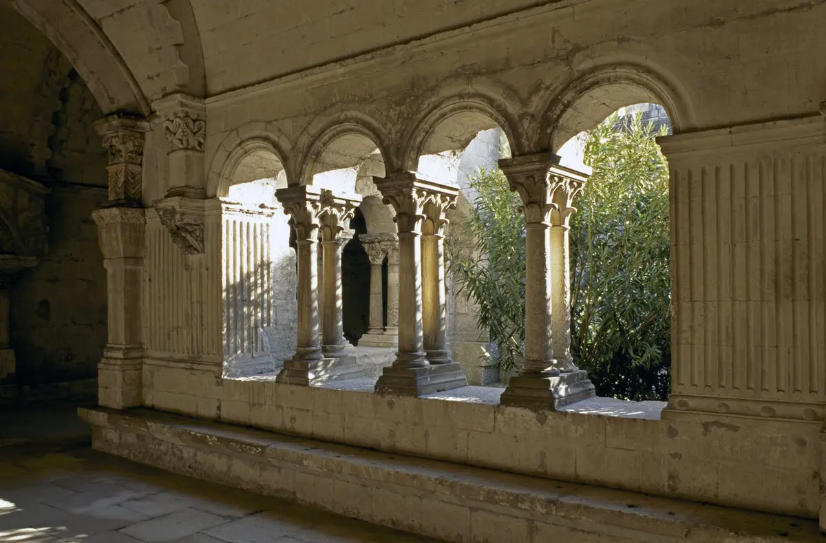 Un couloir en pierre avec des arcs et des colonnes ornés donne sur une cour ensoleillée avec une grande plante feuillue.
