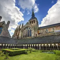 Bâtiment en pierre avec une flèche et des fenêtres cintrées, entouré d'une cour avec des haies taillées et des fleurs épanouies sous un ciel partiellement nuageux. DR