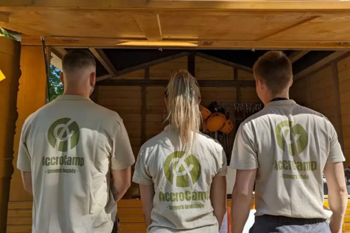 Trois personnes portant des chemises AccroCamp se tiennent face à un hangar en bois ouvert contenant du matériel d'escalade et des casques.