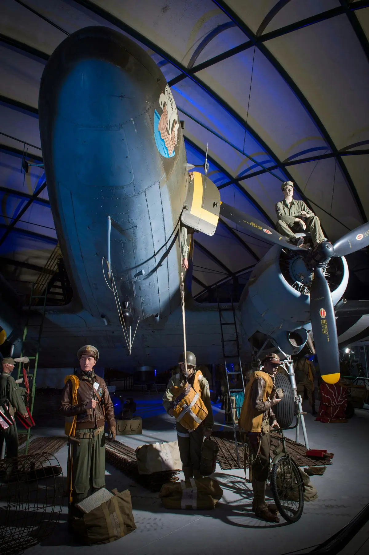 Plusieurs soldats en uniforme de la Seconde Guerre mondiale transportant du matériel sous un grand avion dans un hangar. Un soldat est assis sur le moteur de l'avion.