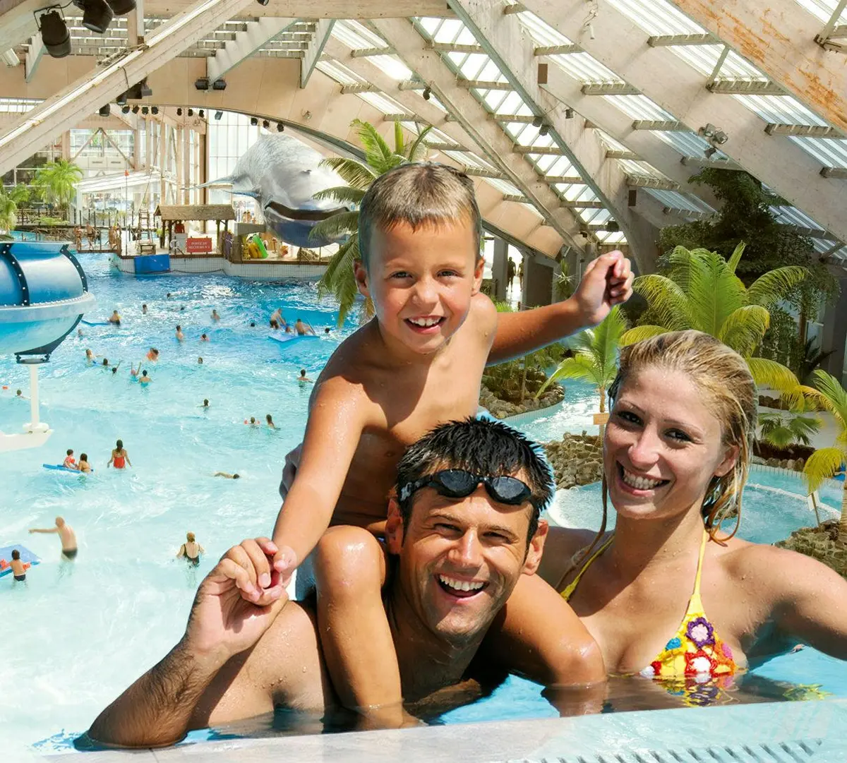 Une famille souriante de trois personnes dans une piscine intérieure au thème tropical, avec un jeune garçon sur les épaules d'un homme et une femme à côté d'eux.