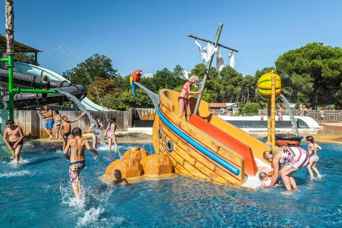 Des enfants s'amusent sur un toboggan aquatique sur le thème d'un bateau pirate, dans une piscine entourée d'arbres et d'autres attractions d'un parc aquatique.