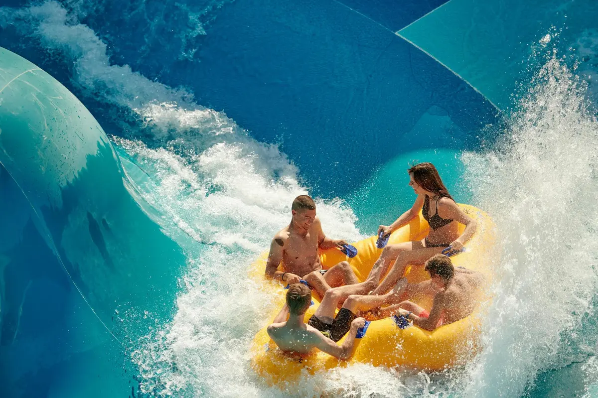 Quatre personnes profitant d'un tour d'eau sur un radeau gonflable jaune, entouré d'eau éclaboussante et d'un toboggan bleu en toile de fond.