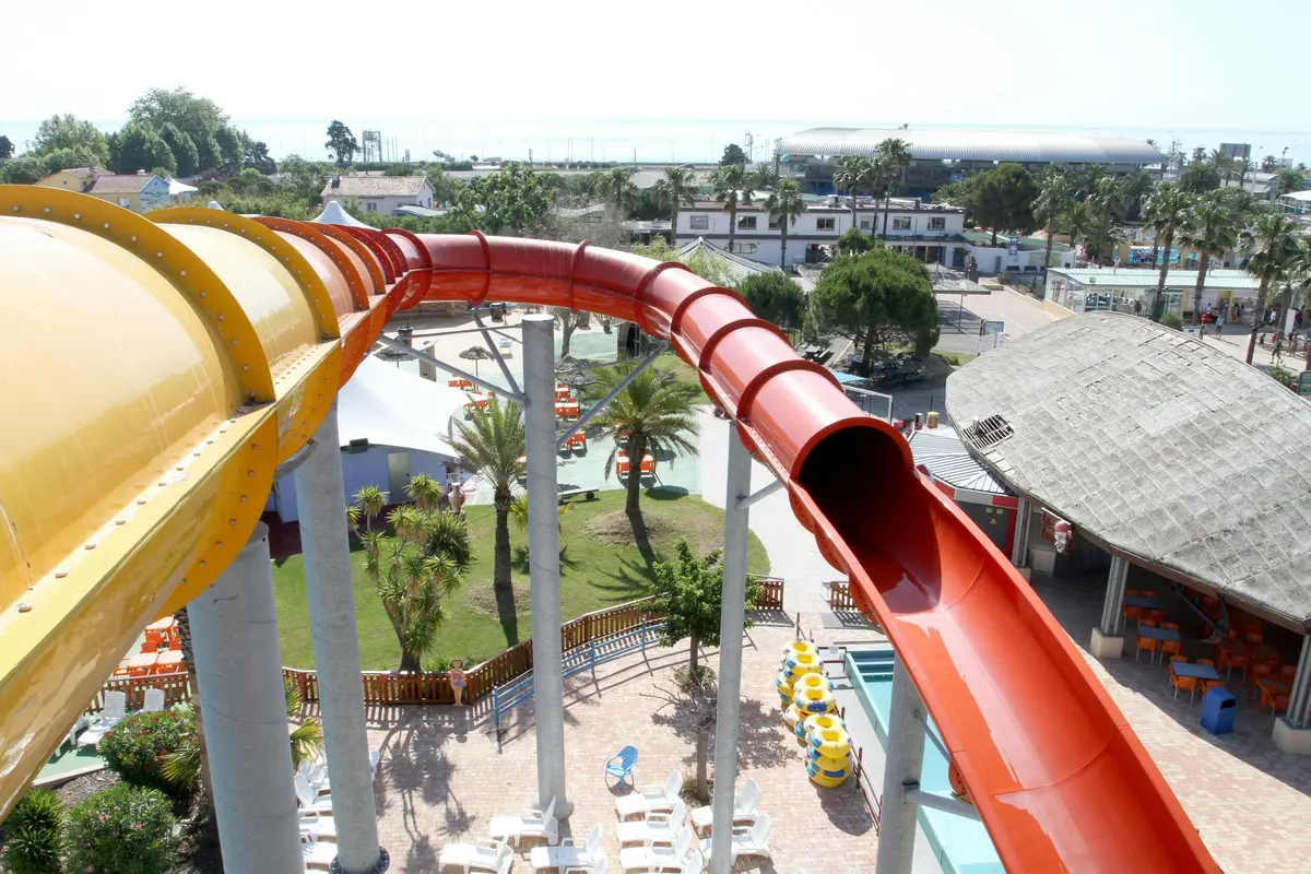 Toboggan aquatique rouge incurvé au-dessus d'une aire de loisirs extérieure ensoleillée avec des palmiers, des bâtiments et des chaises longues.