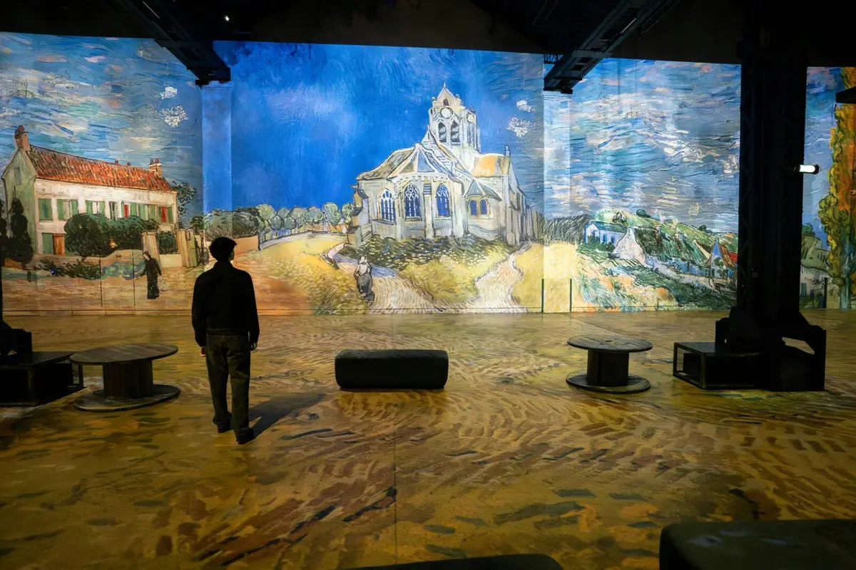 Une personne se tient dans une pièce où sont projetées des œuvres de Van Gogh représentant une église et un paysage, entourée de sièges.