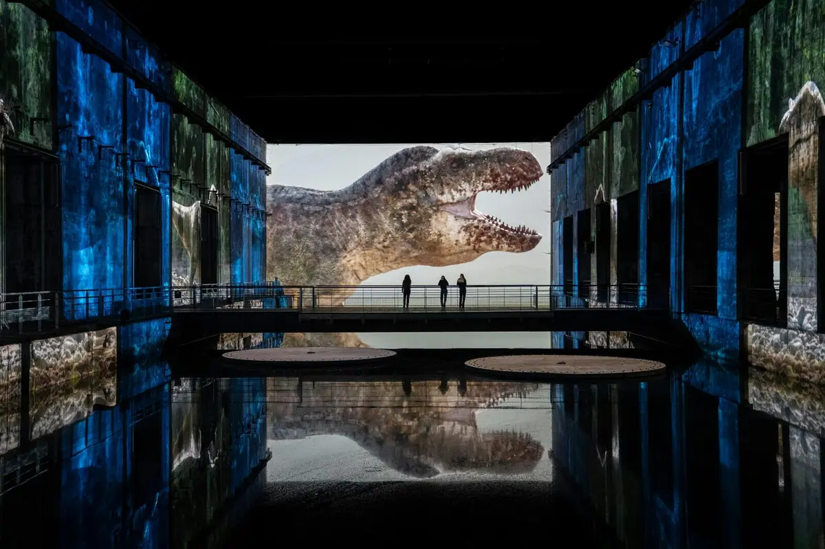 Une grande projection représentant une tête de dinosaure dans une pièce sombre aux murs bleus, avec trois personnes debout sur un pont au centre.