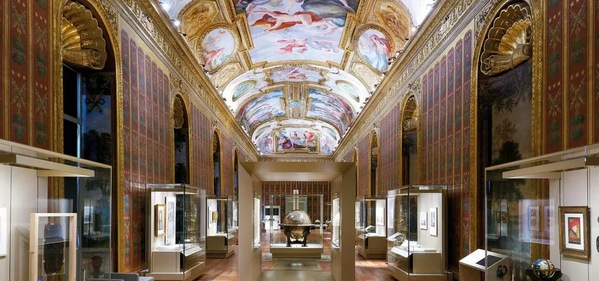 Une galerie richement décorée, avec un plafond peint, des murs ornés et des vitrines abritant des objets et des œuvres d'art.