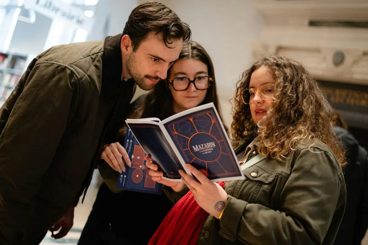 Trois personnes regardent un livret ouvert intitulé Mazarin ; elles semblent en discuter ou l'examiner de près.
