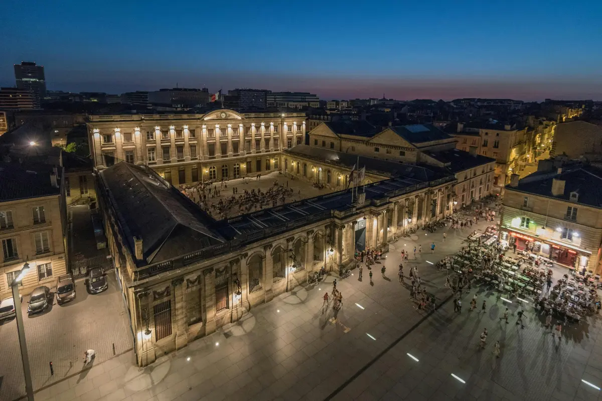 Aerial view of a large courtyard surrounded by historic buildings, with people gathered, illuminated by streetlights at dusk.