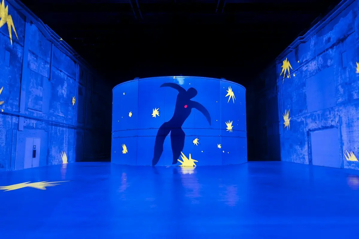 Une installation circulaire bleue comportant une silhouette noire aux traits humains et des formes jaunes ressemblant à des étoiles, dans une pièce sombre.