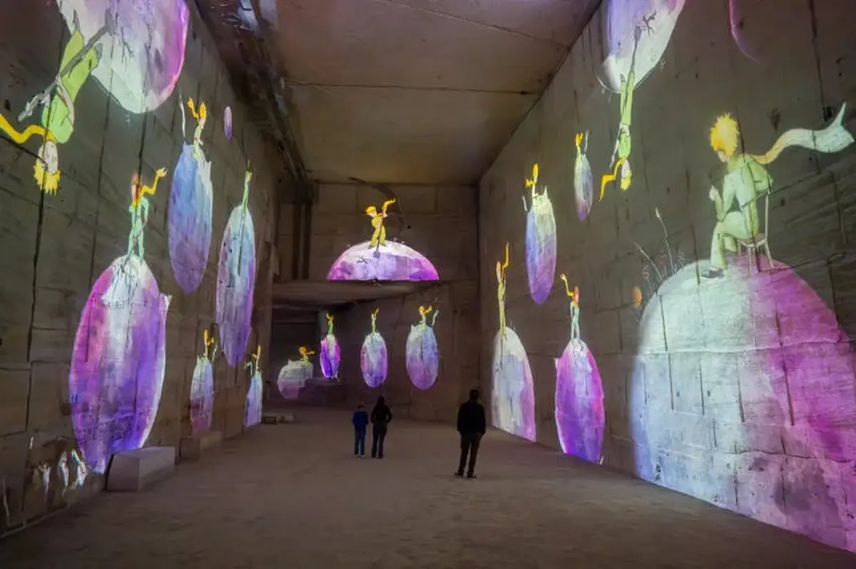 Un grand espace intérieur avec des projections colorées de figures et de planètes sur les murs, et des personnes qui observent l'exposition.