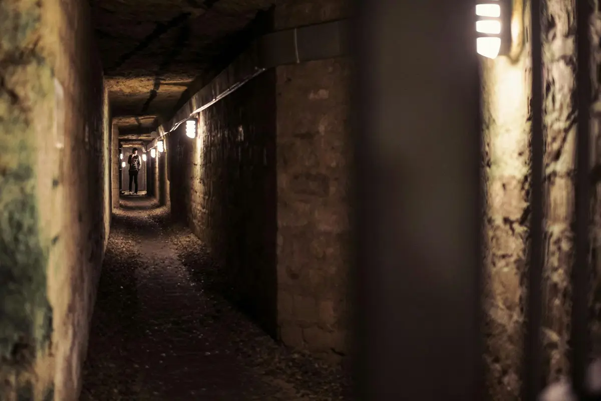 Un tunnel souterrain faiblement éclairé, aux murs bruts, dans lequel on aperçoit au loin une personne marchant sous la lumière des lampadaires.
