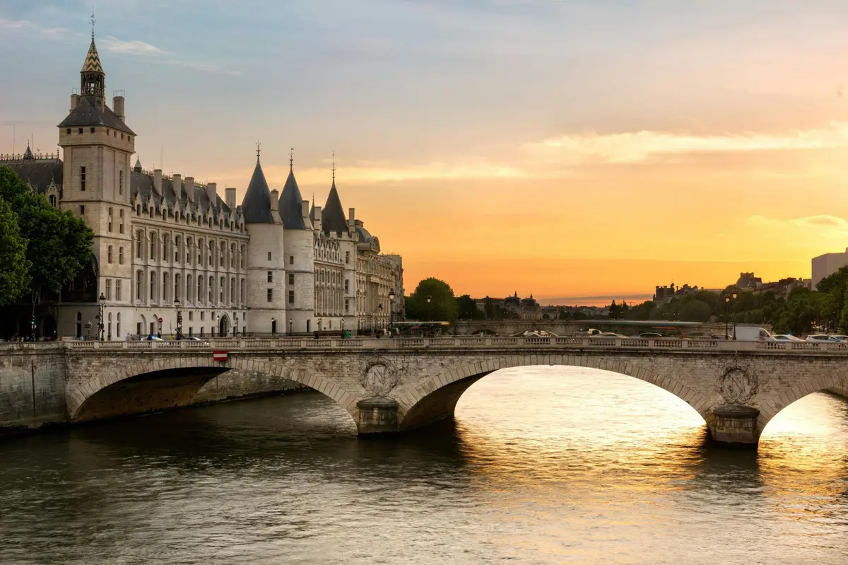 Un bâtiment historique avec des tours au bord d'une rivière, à côté d'un pont en pierre, sous un soleil couchant éclatant.
