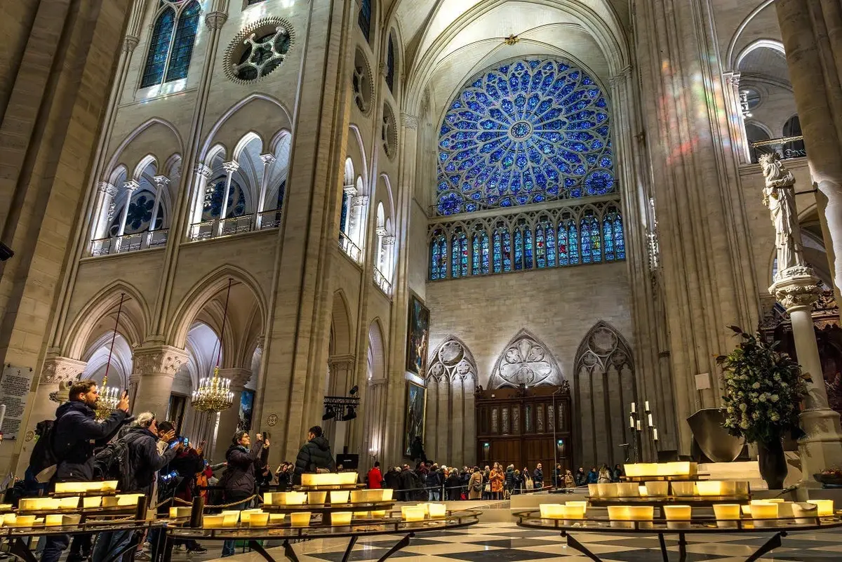 Intérieur d'une cathédrale avec une grande rosace en vitrail bleu, des colonnes voûtées et des visiteurs prenant des photos près de bougies allumées.