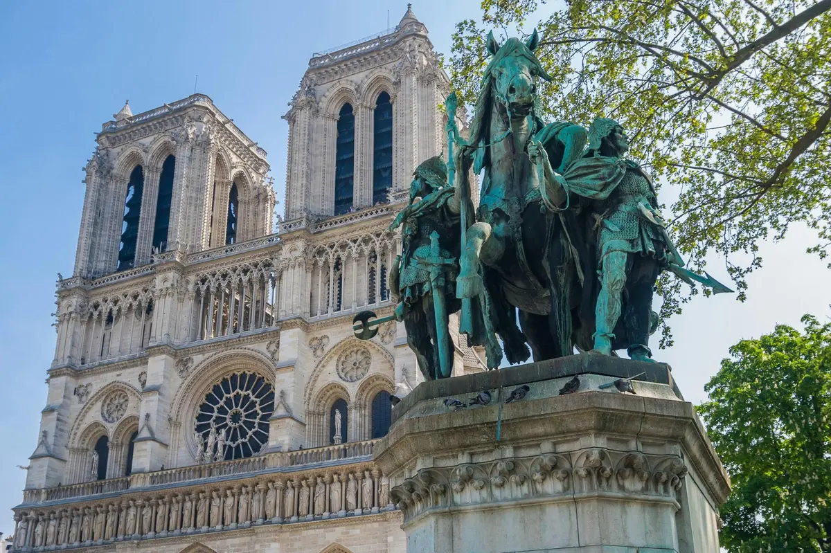 Statue d'un personnage historique à cheval devant la cathédrale Notre-Dame, avec une architecture détaillée et des arbres verdoyants.