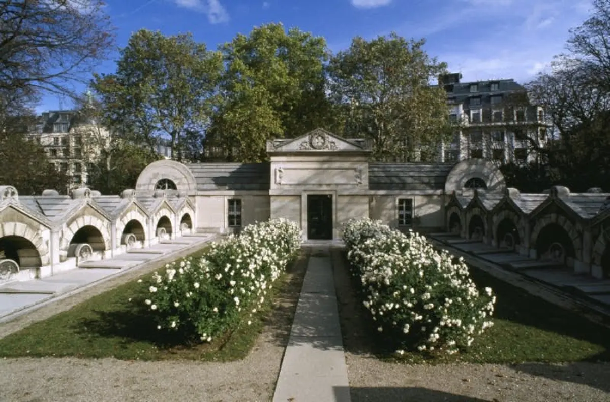 Mausolée en pierre avec des tombes voûtées, entouré de fleurs blanches, d'allées et d'arbres sous un ciel bleu clair.